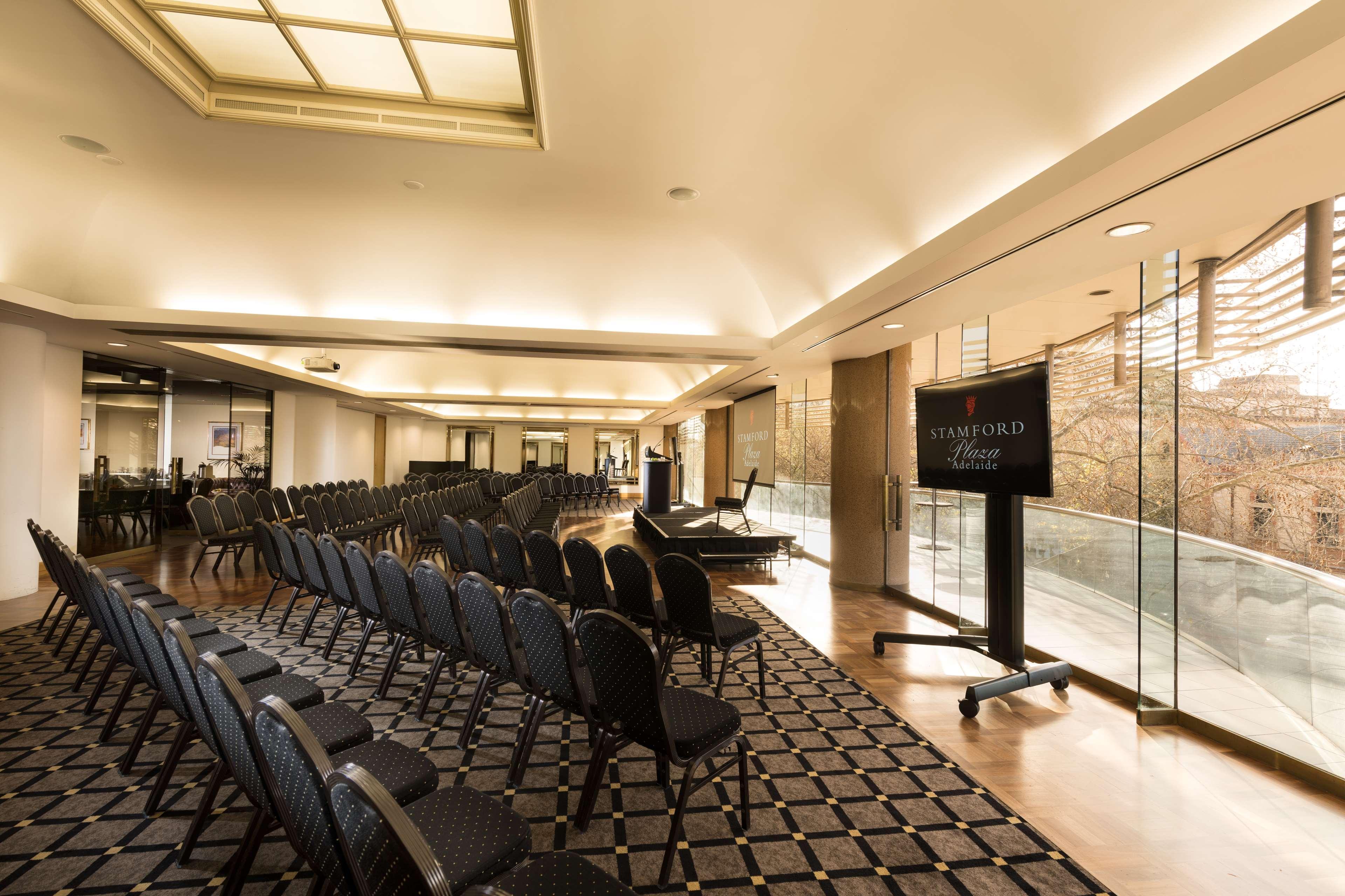Hotel Stamford Plaza Adelaida Exterior foto The photo shows a large, elegantly designed event space or meeting room. There are rows of black chairs set up facing a presentation area. The flooring features a patterned carpet, and large windows allow natural light to enter the room. On one side,